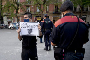 ALBERT SALAMÉ/VILAWEB | Manifestant a la plaça de la Vila de Gràcia de Barcelona amb una pancarta de rebuig que fusiona les paraules Covid i capitalisme, i un Mosso d'Esquadra controlant la seva identitat